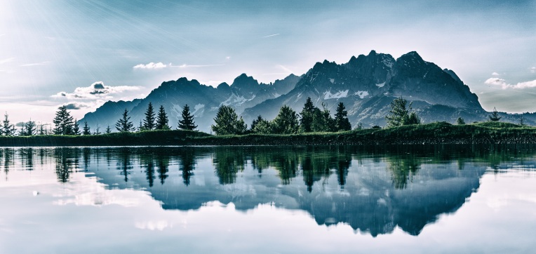 Gray mountains with green trees in front reflected in still waters