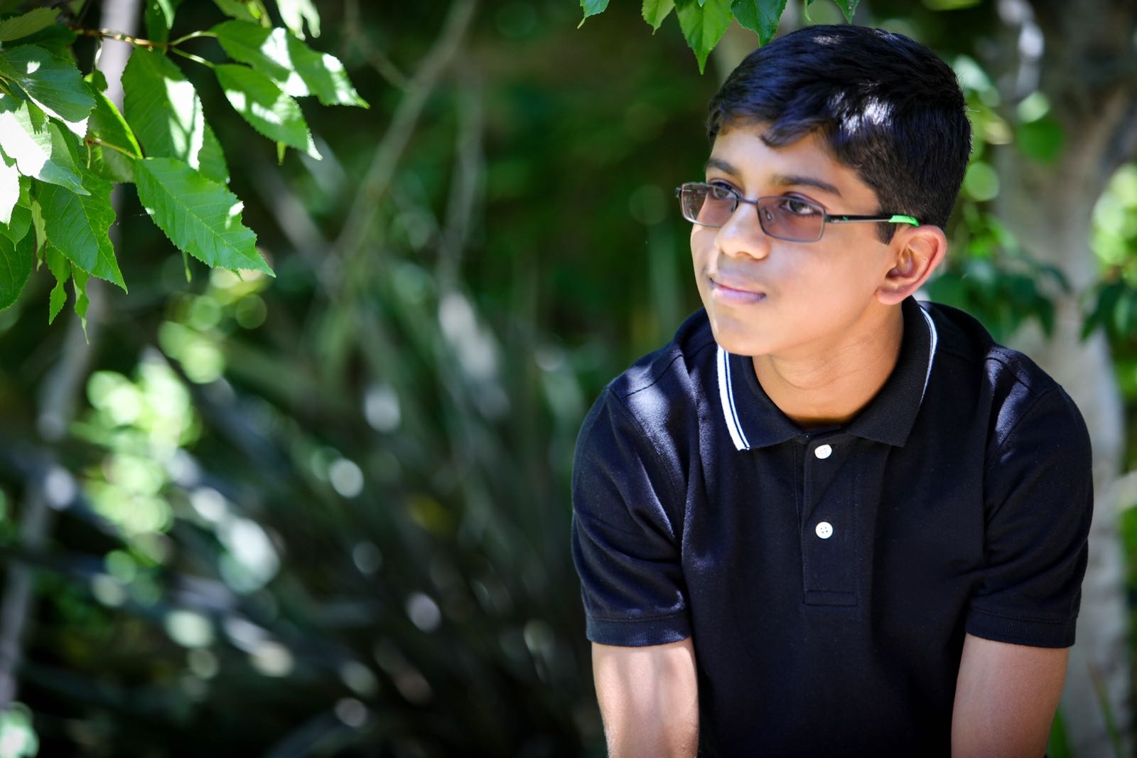 young boy sitting amonst trees wearing a dark shirt and glasses looking off into the distance